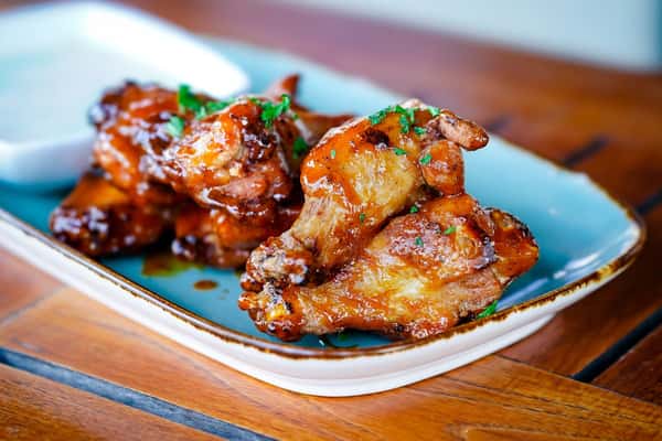 Close-up of crispy glazed chicken wings garnished with chopped herbs, served on a rectangular blue ceramic plate with a side of creamy dipping sauce.