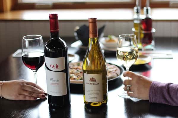 Two diners enjoying red and white wine with artisan pizza at a cozy restaurant table, featuring bottles of Drumheller Chardonnay and Maremma Toscana Rosso.