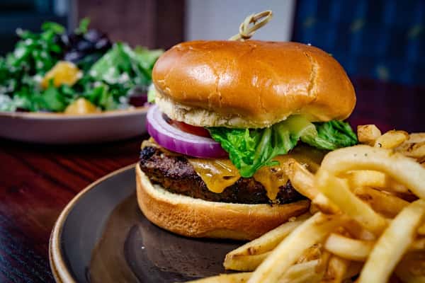 Juicy cheeseburger topped with lettuce, tomato, pickles, and red onion on a toasted bun, served with golden French fries and a side salad in the background.