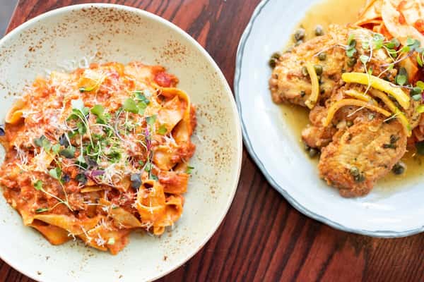 A rustic plate of tagliatelle pasta with tomato meat sauce topped with microgreens and Parmesan, served alongside chicken piccata with lemon slices, capers, and a white wine sauce on a wooden table.