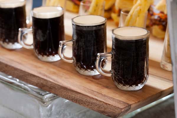 Row of frothy dark beer or root beer served in vintage glass mugs on a wooden tray, accompanied by fries and burgers in the background.