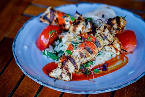 Grilled chicken skewers served over herbed rice with roasted red peppers, microgreens, and a drizzle of vibrant red sauce on a rustic ceramic plate—Mediterranean-inspired restaurant dish.