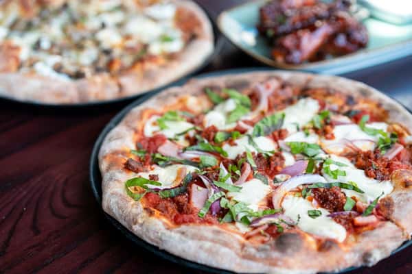 Close-up of a wood-fired pizza topped with fresh mozzarella, red onions, basil, and crumbled sausage, with another pizza and a plate of glazed chicken wings in the background on a dark wooden table.