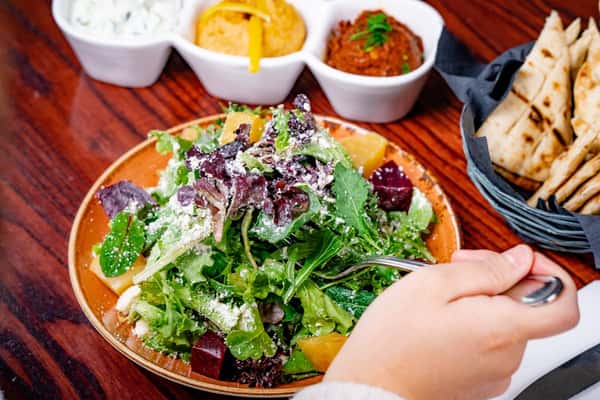 Fresh mixed greens salad with beets and crumbled cheese, accompanied by a trio of Mediterranean dips—tzatziki, hummus, and red pepper spread—with a side of warm pita bread on a dark wooden table.