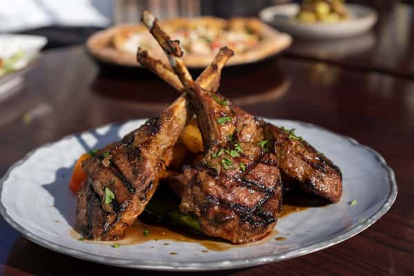 Plate of grilled lamb chops garnished with fresh herbs, served with roasted vegetables, with wood-fired pizza visible in the background on a patio dining table.