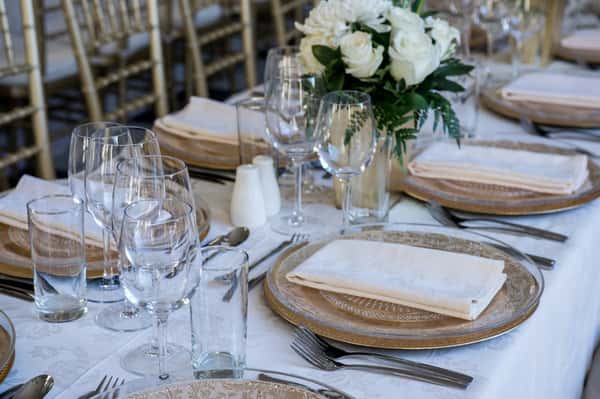 Elegant table setting for a formal event, featuring gold-rimmed chargers, crystal glassware, white linen napkins, and a centerpiece of fresh white roses on a white tablecloth.