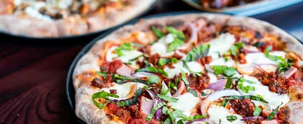 Close-up of a wood-fired pizza loaded with mozzarella, sausage, ham, red onions, and fresh basil on a rustic crust.