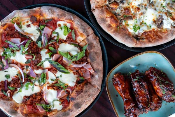 Overhead view of artisan pizzas topped with fresh mozzarella, basil, red onions, and meats, paired with saucy glazed chicken wings on a ceramic plate.