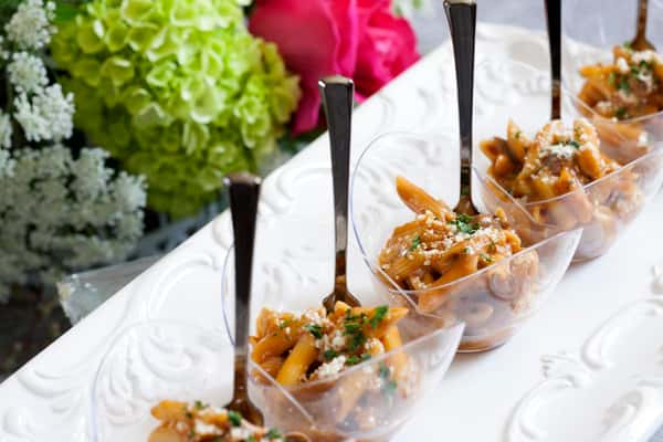 Elegant appetizer presentation of mini penne pasta cups in creamy tomato sauce, garnished with grated cheese and fresh herbs, served with black mini forks on a decorative white tray beside a floral arrangement.