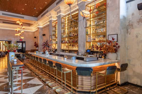 Elegant bar interior featuring a long marble counter, gold-accented bar stools, floral arrangements, and backlit liquor shelves.