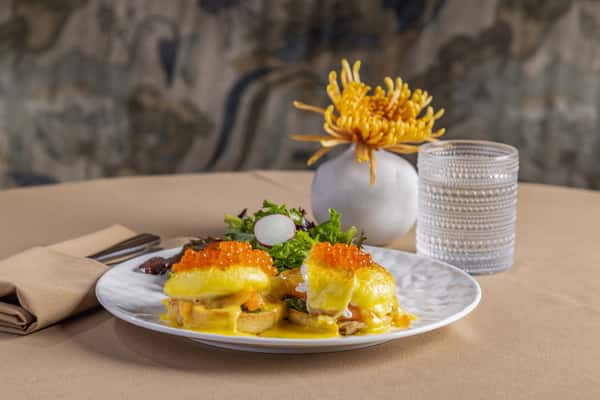 Eggs Benedict with hollandaise sauce and salmon roe, served with a fresh salad on a white plate with elegant table setting.
