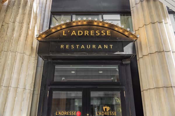 Black and gold entrance sign of L'Adresse Restaurant framed by tall stone columns and decorative light bulbs.