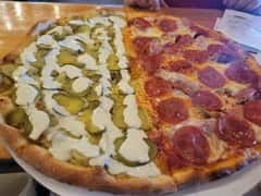 Half pepperoni pizza and half pickle pizza with creamy drizzle, served on a round tray in a casual dining setting.