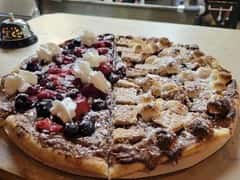 Half dessert pizza topped with whipped cream and mixed berries, and half with marshmallows, cookies, and powdered sugar on chocolate spread.