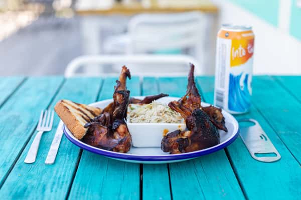 The image features grilled lamb chops served over creamy mashed potatoes, garnished with fresh herbs. Plated on a rustic wooden board atop a bright turquoise table, the dish has a vibrant and upscale presentation.