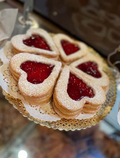 A tray of delicate heart-shaped cookies is beautifully arranged and dusted with powdered sugar. Each cookie is filled with vibrant red jam in the center, creating a charming and sweet treat that's perfect for dessert or a bakery display.