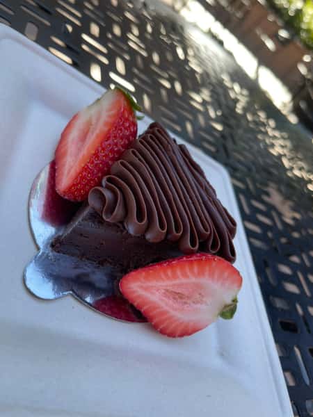 A rich slice of chocolate cake is topped with smooth chocolate frosting and garnished with fresh strawberry slices. The dessert sits on a white plate, creating a beautiful contrast between the deep chocolate layers and the bright red fruit, making it look indulgent and elegant.