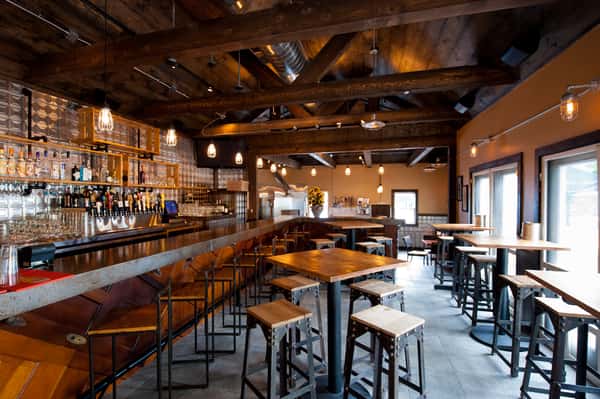 Rustic industrial restaurant interior with wooden beams, bar seating, high-top tables, and warm ambient lighting