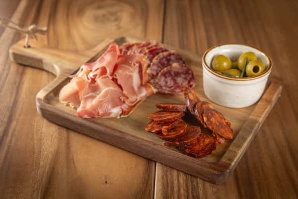 A wooden board displaying cured meats (prosciutto, chorizo) and a small bowl of olives.