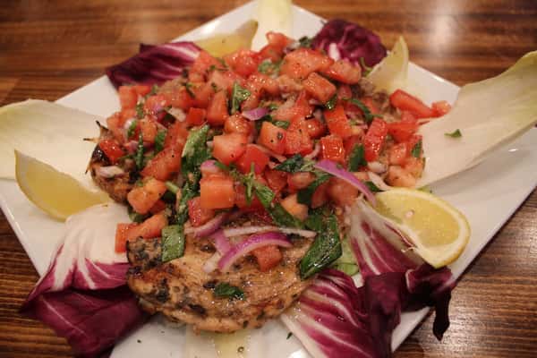 Grilled chicken topped with a vibrant bruschetta mix of diced tomatoes, red onions, basil, and olive oil, surrounded by endive leaves and lemon wedges.