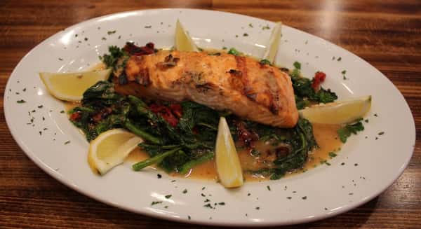 Grilled salmon fillet served over sautéed spinach and sun-dried tomatoes, garnished with lemon wedges and fresh herbs on a white plate.