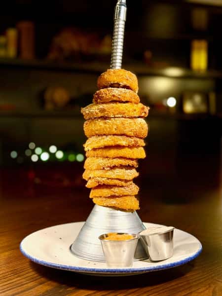 Crispy golden onion rings stacked high on a metal tower stand, served with dipping sauces on a round white plate.