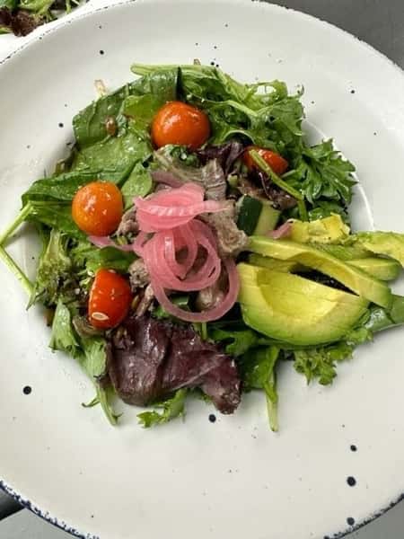 Mixed greens salad with cherry tomatoes, avocado slices, pickled red onions, cucumber, and a light vinaigrette dressing, served in a rustic white bowl.