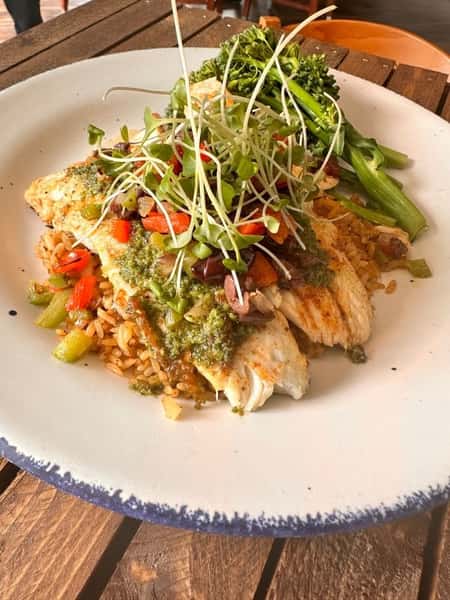Grilled fish fillet over seasoned rice, topped with chimichurri, roasted peppers, olives, and microgreens, with a side of broccolini.