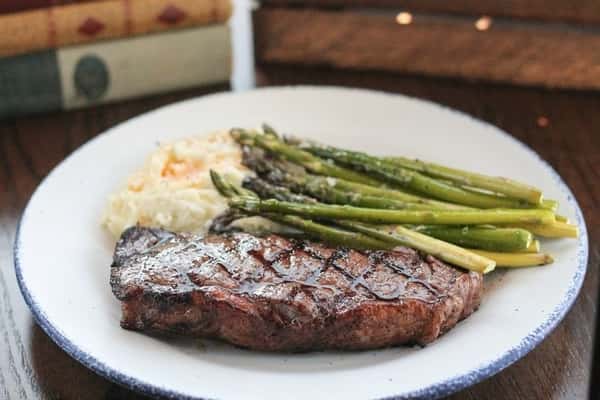 Grilled New York strip steak with perfect sear marks, served with roasted asparagus and cheesy mashed potatoes on a rustic white plate.