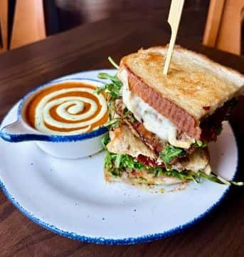 Hearty grilled sandwich layered with greens, cheese, and bacon, served alongside creamy tomato soup with a decorative swirl of cream in a rustic bowl.