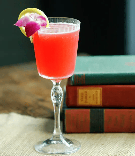 Bright pink cocktail garnished with a lemon wheel and purple orchid flower, served in a vintage coupe glass on a rustic wooden table with stacked books in the background.