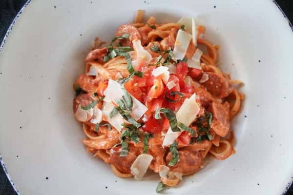 Creamy tomato-based pasta topped with sliced Italian sausage, fresh basil, diced tomatoes, and shaved Parmesan cheese, served in a rustic white bowl.