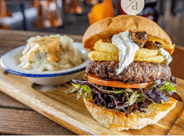 Juicy gourmet burger with lettuce, tomato, fried onion rings, mushrooms, and creamy sauce on a brioche bun, paired with mashed potatoes topped with gravy.