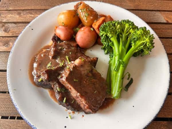 Slow-braised beef topped with rich gravy, served with baby red potatoes, carrots, and steamed broccolini on a rustic white plate.