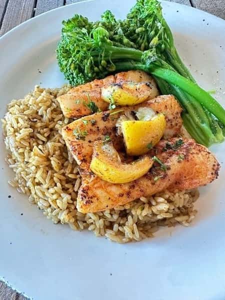 Grilled white fish fillets topped with lemon wedges, served with seasoned rice pilaf and steamed broccolini on a wide white plate.