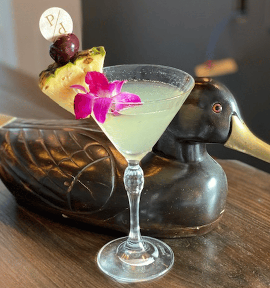 Refreshing martini-style cocktail garnished with pineapple, a dark cherry, and a vibrant orchid flower, served in a stemmed glass next to a decorative duck sculpture.