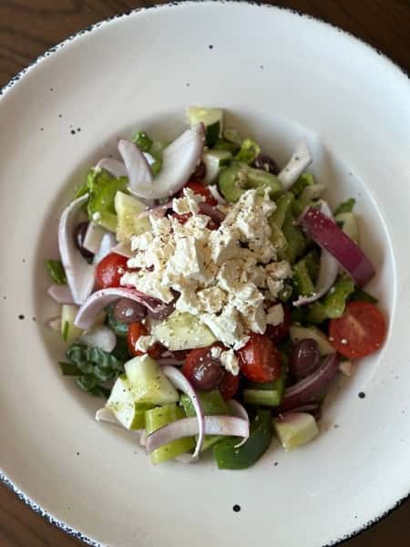 Fresh Greek salad with chopped cucumbers, cherry tomatoes, red onions, Kalamata olives, green bell peppers, and crumbled feta cheese, served in a wide white ceramic bowl.