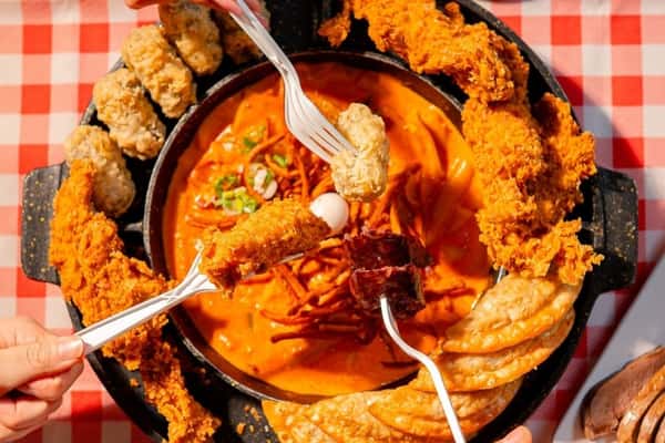 Hands holding forks with fried foods dipping into a spicy orange tteokbokki surrounded by crispy chicken and dumplings.