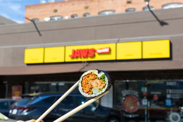 A hand holding chopsticks with a gimbap roll in focus outside Jaws TPK restaurant with a bright yellow storefront sign.