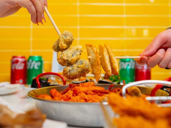 Hands holding skewers of fried dumplings and tempura over a pan of tteokbokki with colorful soda cans in the background.