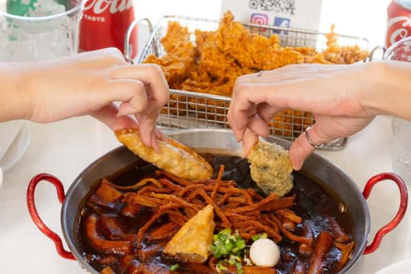 Two hands dipping fried dumplings into a pan of dark spicy tteokbokki, with fried snacks and soda cans in the background.