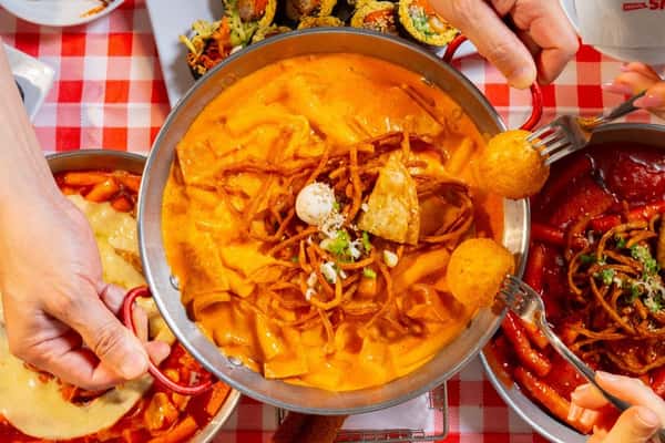 Hands holding forks and bowls of creamy, spicy tteokbokki surrounded by kimbap on a red checkered tablecloth.