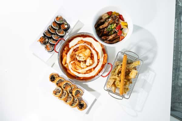 Assorted Korean dishes including cheesy tteokbokki, gimbap, bulgogi rice bowl, and vegetable tempura on a white table.