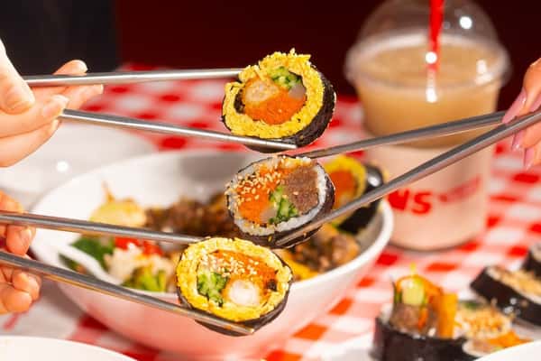 Three hands holding chopsticks with colorful kimbap pieces over a red checkered table with bowls and a drink.