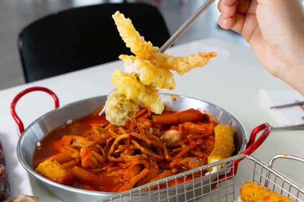 Chopsticks holding assorted tempura above a pan of spicy Korean tteokbokki topped with fried noodles and fish cakes.