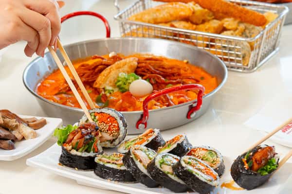 Chopsticks picking up a gimbap roll with tteokbokki and assorted fried snacks in the background on a white table.