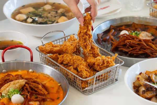 A hand picking up crispy fried chicken from a basket surrounded by Korean dishes like tteokbokki and soup bowls.