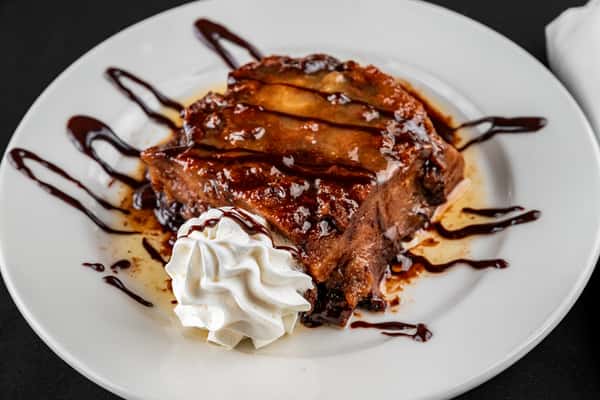 Decadent slice of Southern-style bread pudding drizzled with rich chocolate sauce and caramel glaze, served with a swirl of whipped cream on a white dessert plate.