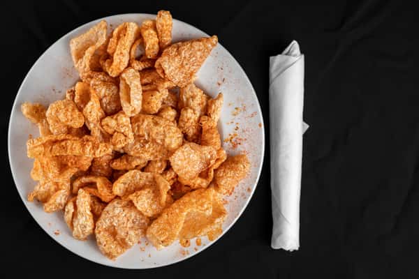 Crispy seasoned pork rinds piled high on a white plate, lightly dusted with spicy paprika for a bold Southern-style snack.