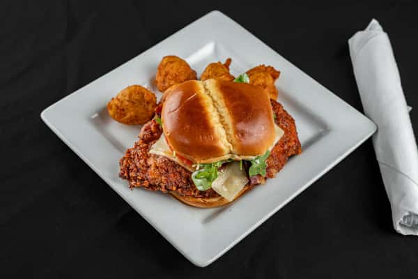 Crispy fried chicken sandwich with lettuce, tomato, cheese, and a toasted brioche bun, served with golden hushpuppies on a square white plate.
